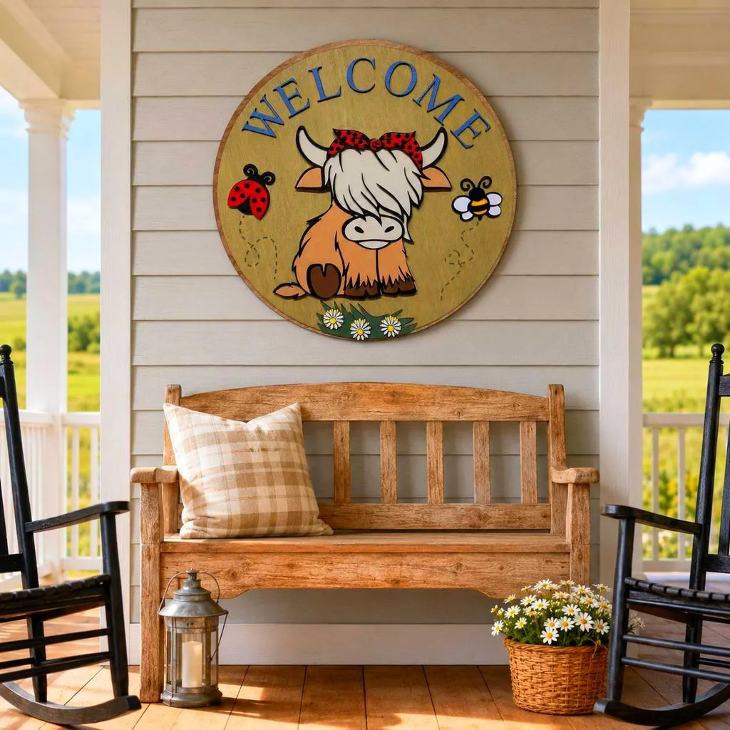 Highland cow Welcome wood sign displayed above a rustic bench between black rocking chairs on a sunny farmhouse porch.