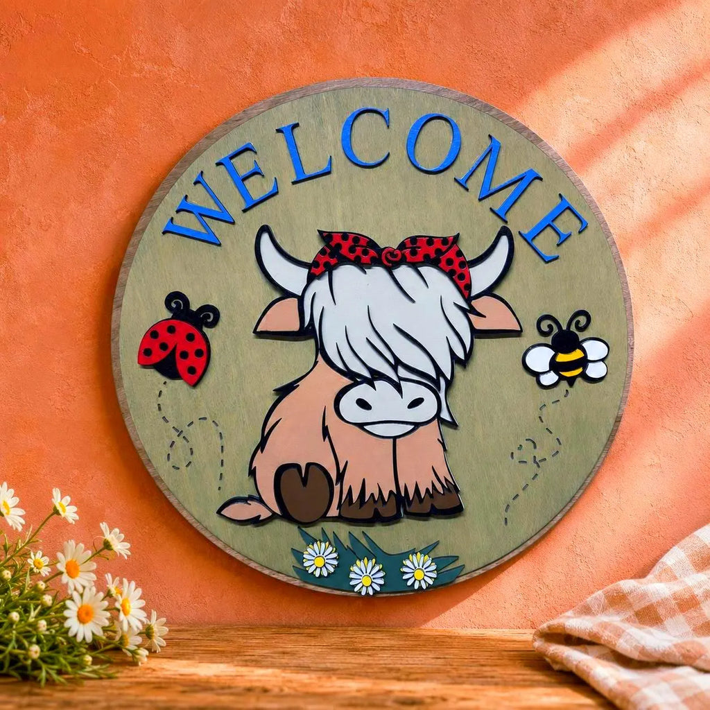 Close-up of a layered Highland cow Welcome sign against a peach textured background with daisies and neutral gingham fabric.