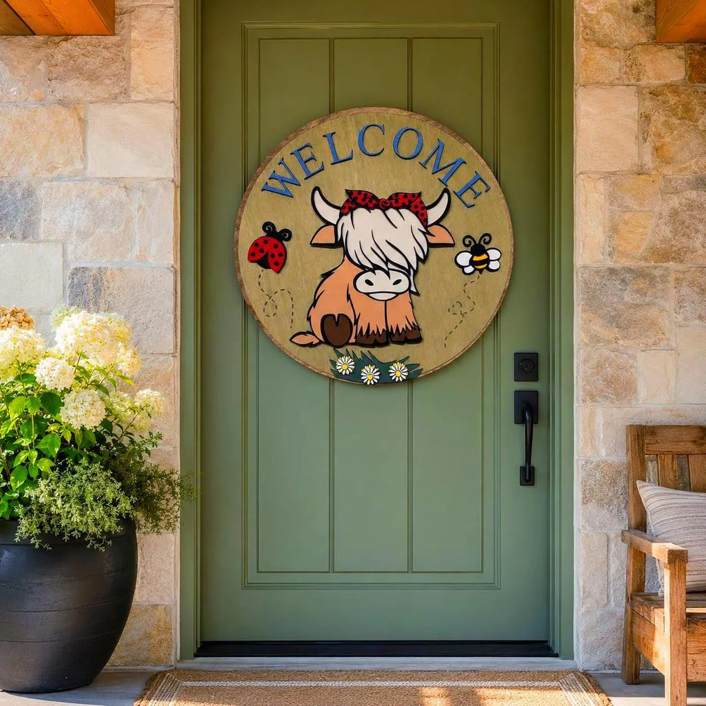 Round layered wood Welcome sign with a seated Highland cow, red polka-dot bow, ladybug, bee and daisies on a sage farmhouse door.