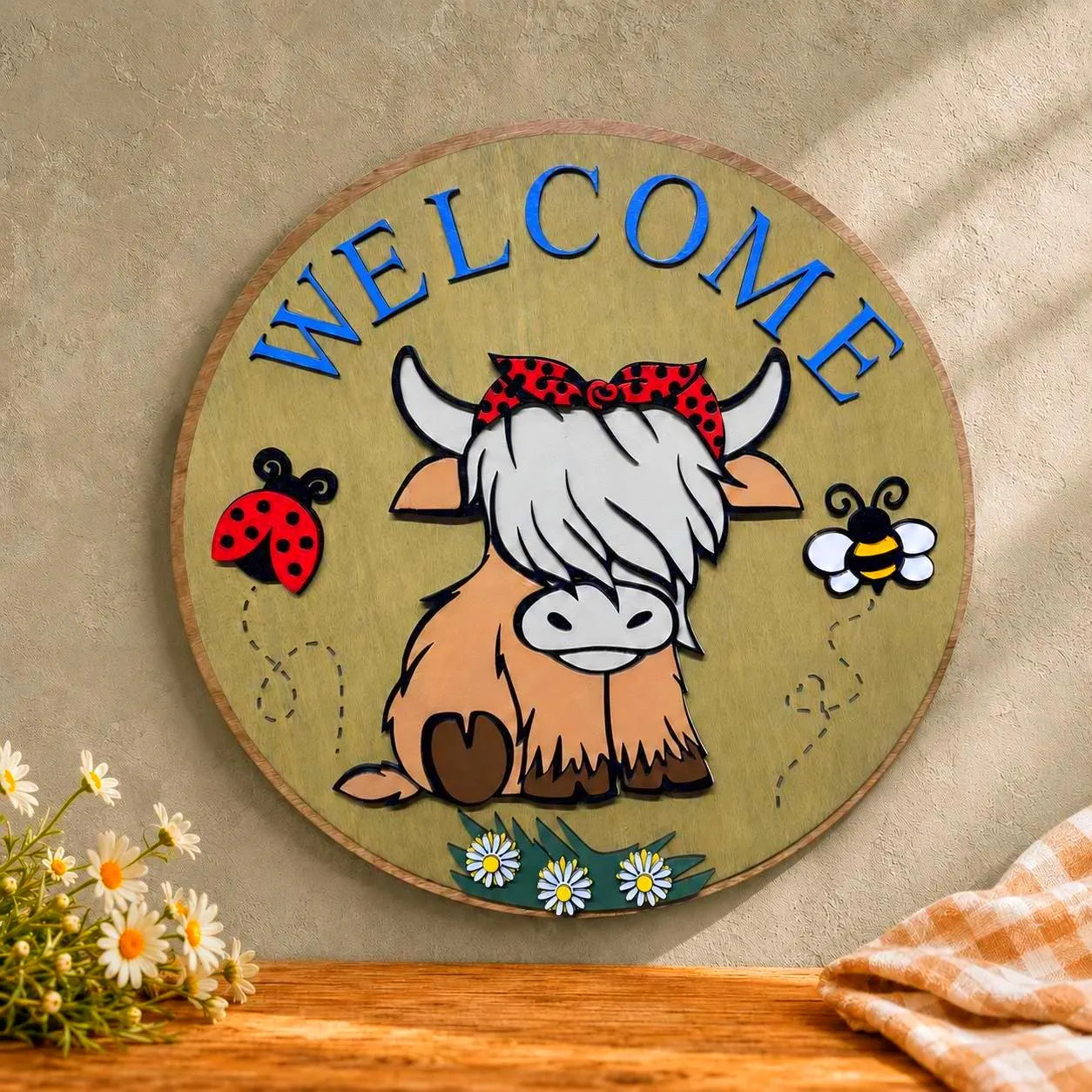 Close-up of a round Highland cow Welcome wood sign against a warm beige wall with daisies and folded gingham fabric.